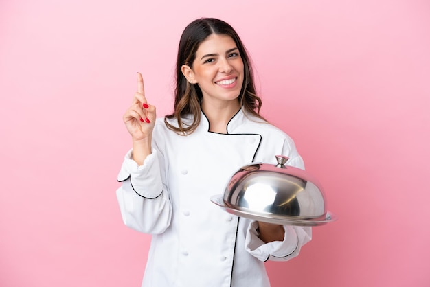 Joven chef italiana sosteniendo una bandeja con tapa aislada en un fondo rosado señalando una gran idea