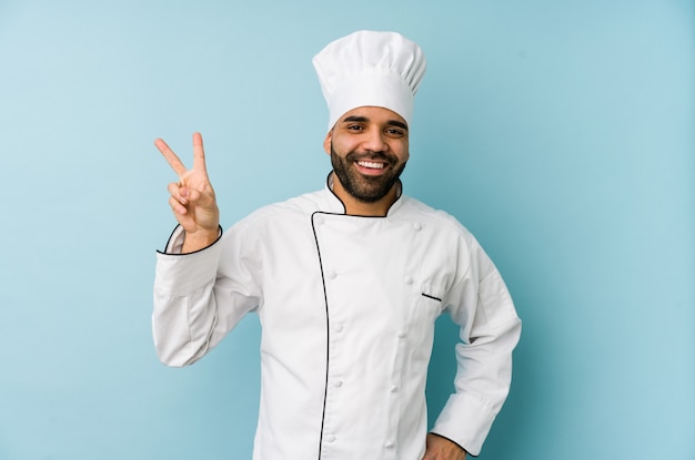 Joven chef latino hombre alegre y despreocupado mostrando un símbolo de paz con los dedos.