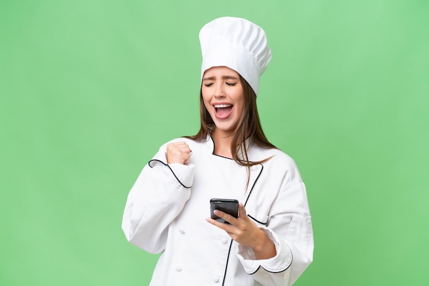 Joven chef mujer caucásica sobre fondo aislado con teléfono en posición de victoria