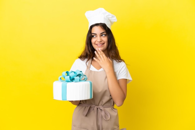 Joven chef pastelera caucásica con un gran pastel aislado de fondo amarillo mirando hacia arriba mientras sonríe