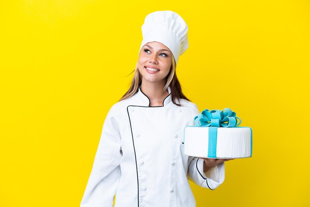 Joven chef pastelera rusa con un gran pastel aislado en un fondo amarillo pensando en una idea mientras mira hacia arriba