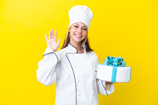 Joven chef pastelera rusa con un gran pastel aislado de fondo amarillo saludando con la mano con expresión feliz