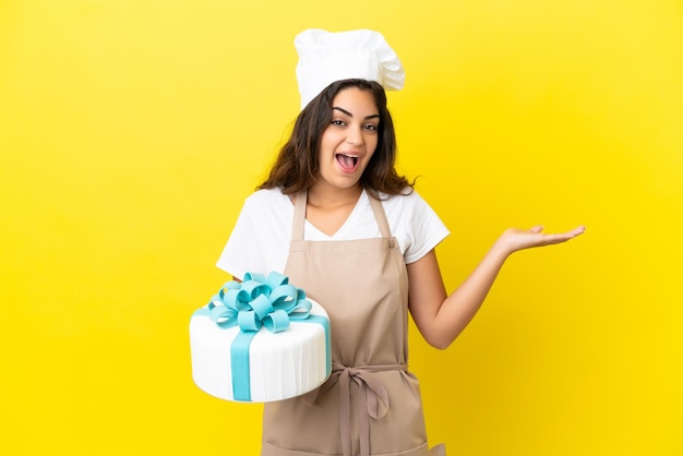 Joven chef de pastelería caucásica con un gran pastel aislado de fondo amarillo con expresión facial sorprendida