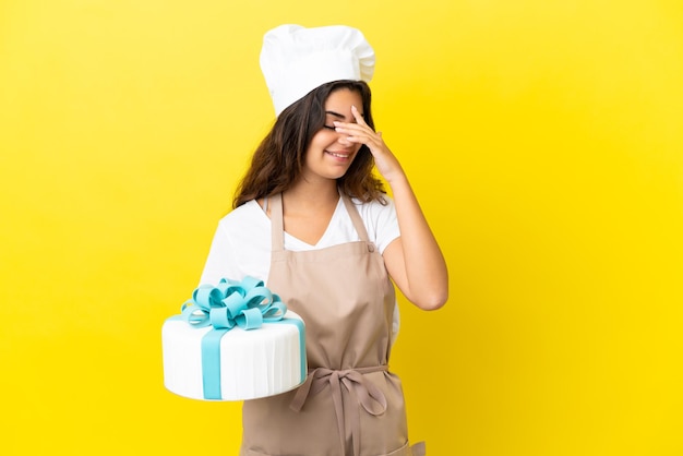 Joven chef de pastelería caucásica con un gran pastel aislado de fondo amarillo riendo