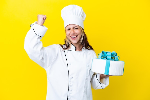 Joven chef de pastelería rusa con un gran pastel aislado de fondo amarillo celebrando una victoria