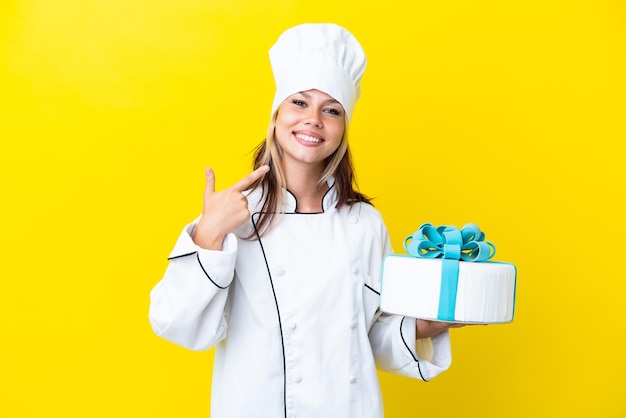 Joven chef de pastelería rusa con un gran pastel aislado de fondo amarillo dando un gesto de pulgar hacia arriba