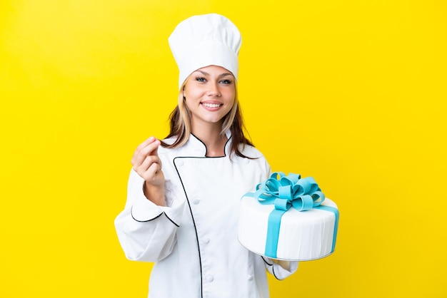 Joven chef de pastelería rusa con un gran pastel aislado de fondo amarillo haciendo gestos de dinero