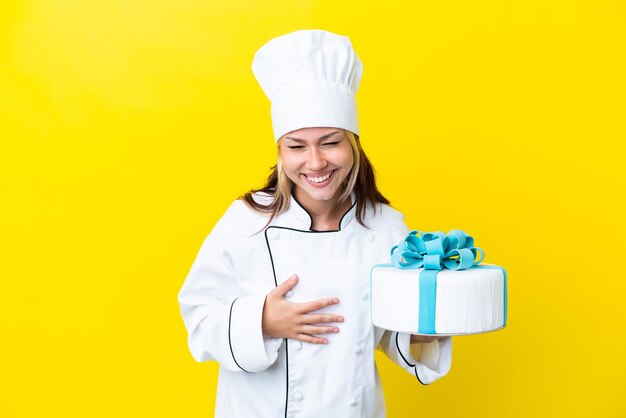 Joven chef de pastelería rusa con un gran pastel aislado de fondo amarillo sonriendo mucho