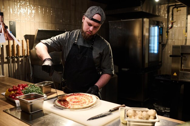 Joven chef profesional en uniforme esparciendo ingredientes en pan de pizza