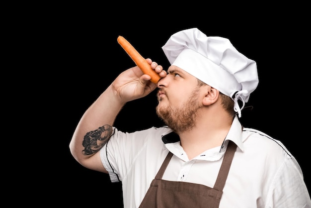 Un joven chef serio con sombrero y delantal sosteniendo zanahoria cruda cerca de la nariz y mirando hacia otro lado