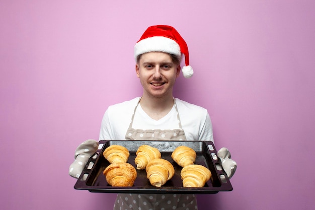 joven chef en sombrero de Santa y delantal sostiene hoja de hornear con croissants