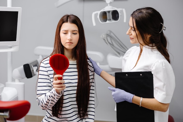 Joven chica bonita mirándose tristemente en el espejo mientras está sentada en la silla dental y la doctora está alentando al paciente sosteniendo su hombro