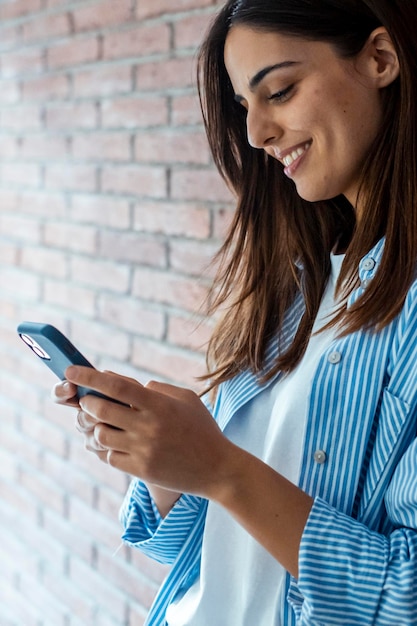 Joven chica caucásica moderna sonriendo mira el teléfono móvil de pie apoyándose en la pared de ladrillo mujer relajada mira el teléfono concepto de tecnología