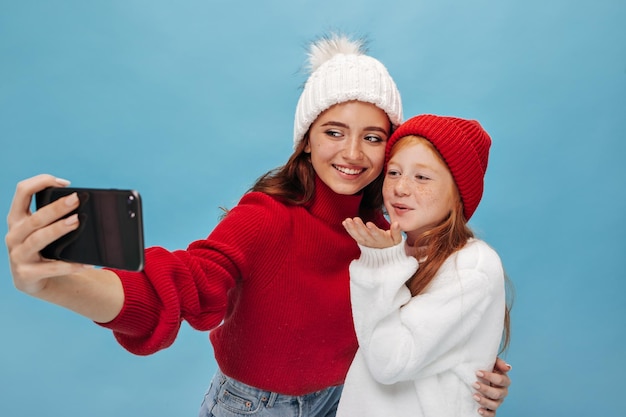 Joven chica moderna con pelo de jengibre y pecas sopla un beso y posa con una hermosa hermana sonriente con sombrero blanco en un fondo aislado