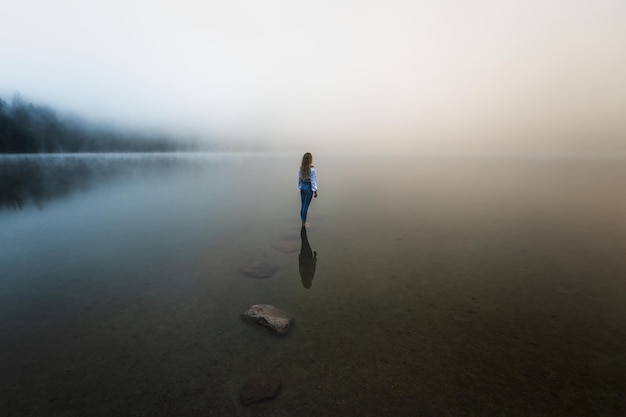 Joven chica rubia de pie en el lago de niebla al amanecer en el bosque salvaje