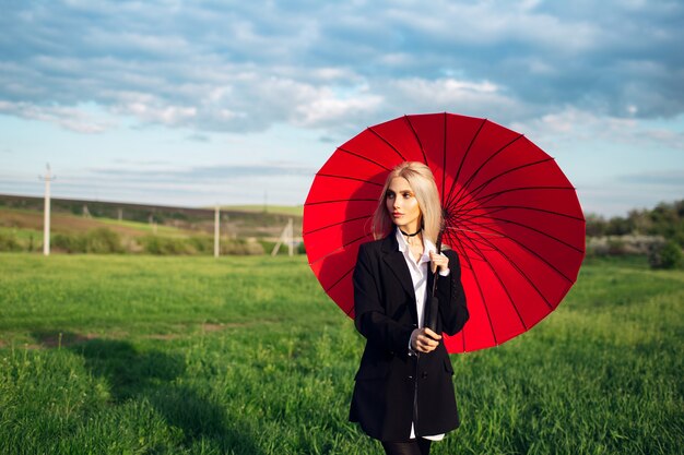 Joven chica rubia sosteniendo paraguas rojo sobre fondo de campo verde y azul cielo nublado