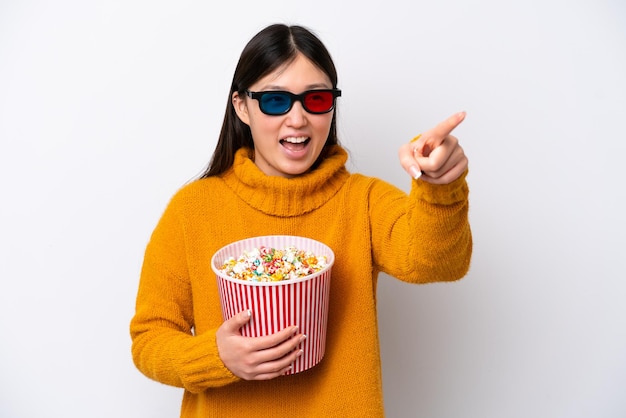 Joven china aislada de fondo blanco con gafas 3d y sosteniendo un gran cubo de palomitas de maíz mientras señala hacia otro lado