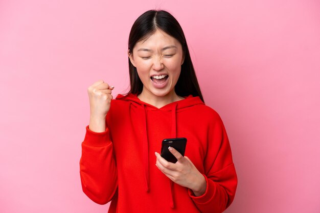 Joven china aislada de fondo rosa usando teléfono móvil y haciendo gesto de victoria