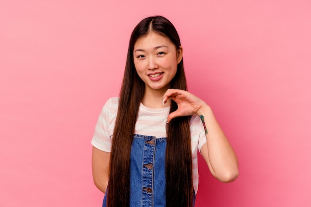 Joven China aislada en la pared rosa sonriendo y mostrando una forma de corazón con las manos.