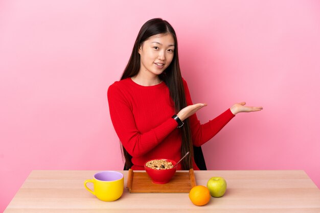 Joven china desayunando en una mesa extendiendo las manos a un lado para invitar a venir