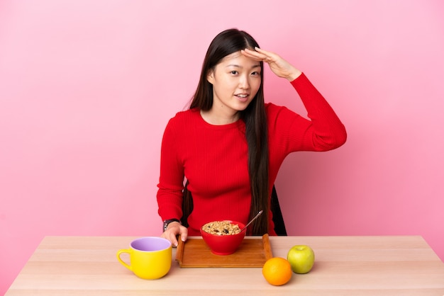 Joven china desayunando en una mesa mirando lejos con la mano para mirar algo