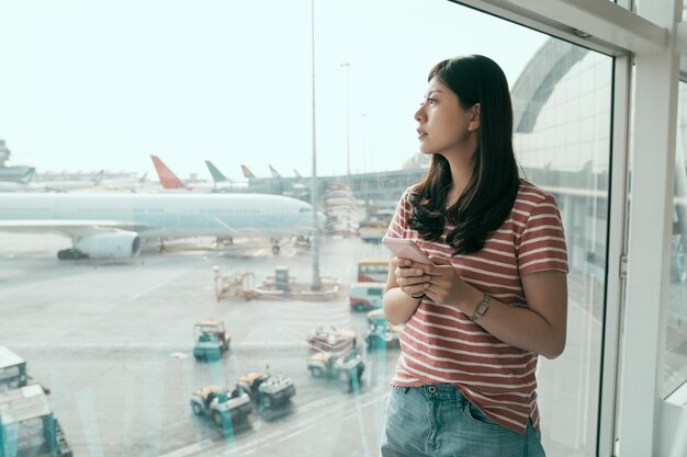 Joven china está parada cerca de la ventana en el aeropuerto y mirando el avión antes de la salida. chica asiática en ropa casual viajes vacaciones de verano. mujer viajera utiliza teléfono móvil comprobar horario de vuelo