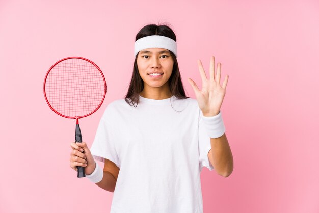 Joven china jugando bádminton en una pared rosa sonriendo alegre mostrando el número cinco con los dedos.