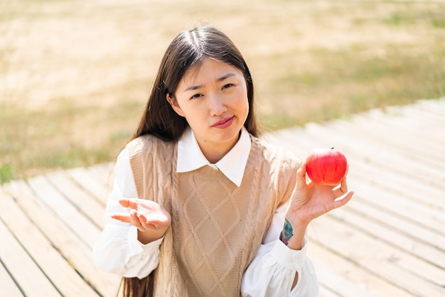 Joven china con una manzana al aire libre haciendo gesto de dudas mientras levanta los hombros