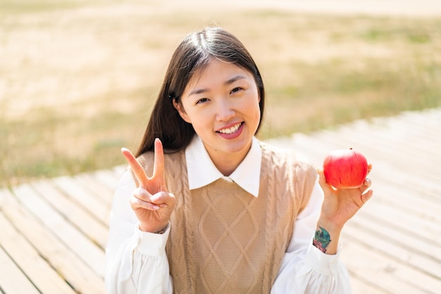 Joven china con una manzana al aire libre sonriendo y mostrando el signo de la victoria