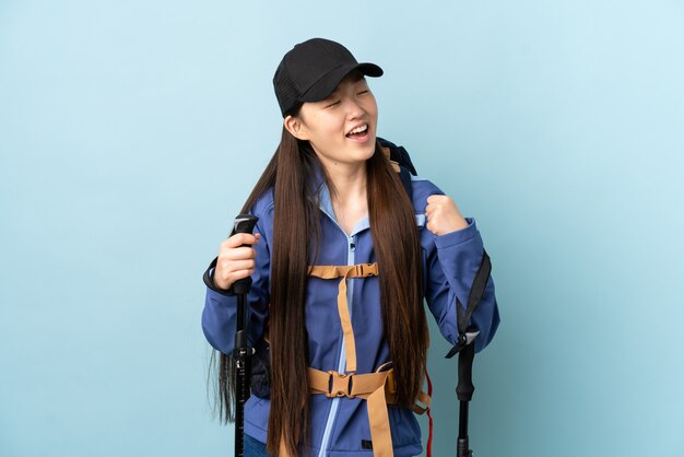 Joven china con mochila y bastones de trekking sobre pared azul aislado celebrando una victoria