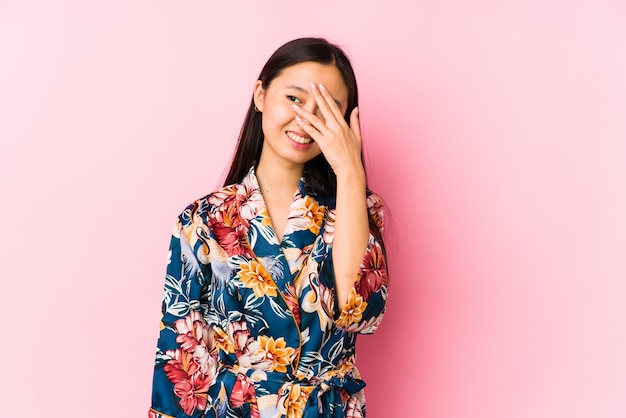 Joven china con un pijama de kimono aislado parpadeo a través de los dedos, avergonzado cubriendo la cara.