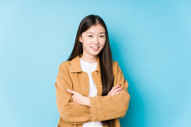 Joven china posando en una pared azul aislada que se siente segura, cruzando los brazos con determinación.