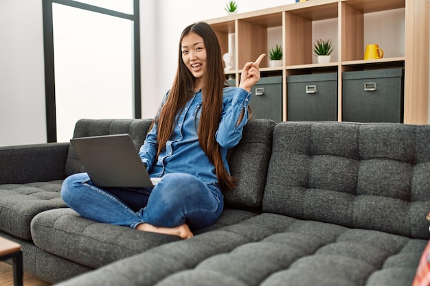 Joven china con ropa informal trabajando con una laptop en casa sonriendo feliz señalando con la mano y el dedo a un lado