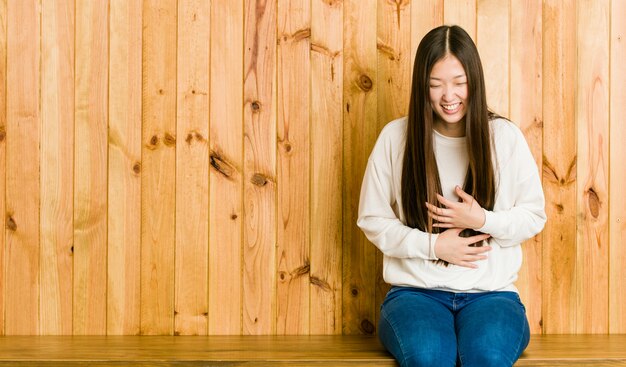 Joven china sentada en un lugar de madera se ríe alegremente y se divierte manteniendo las manos sobre el estómago.