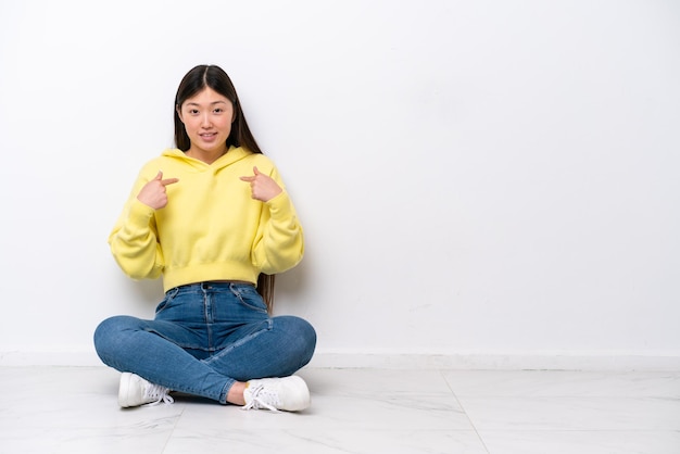 Joven china sentada en el suelo aislada en la pared blanca con expresión facial sorpresa