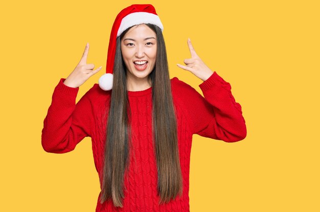 Joven china con sombrero de navidad gritando con expresión loca haciendo símbolo de rock con las manos arriba. estrella de la música concepto de música pesada.