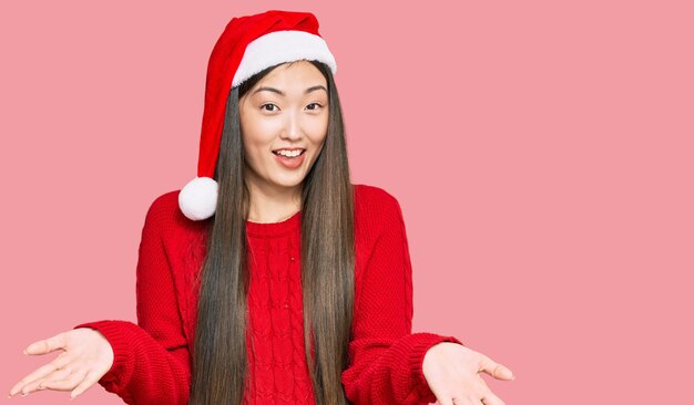Joven china con sombrero de navidad sonriendo alegre ofreciendo manos dando asistencia y aceptación