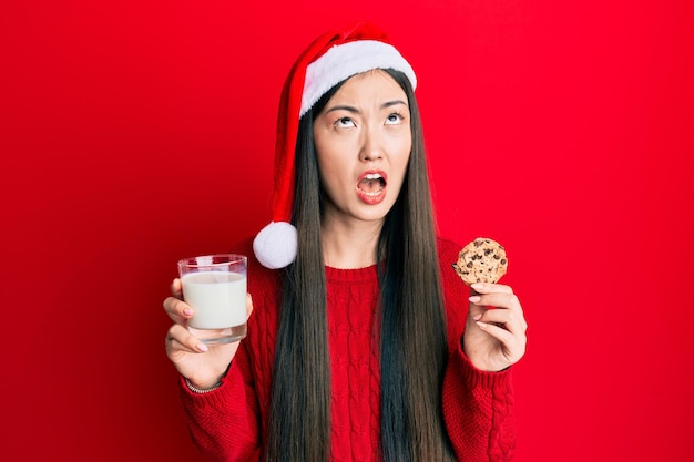 Joven china con sombrero de navidad sosteniendo galletas y leche enojada y loca gritando frustrada y furiosa, gritando con ira mirando hacia arriba.