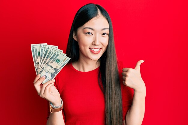 Joven china sosteniendo un billete de 20 dólares sonriendo feliz y positiva, pulgar arriba haciendo excelente y señal de aprobación