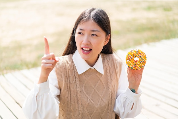 Joven china sosteniendo un donut al aire libre con la intención de darse cuenta de la solución mientras levanta un dedo