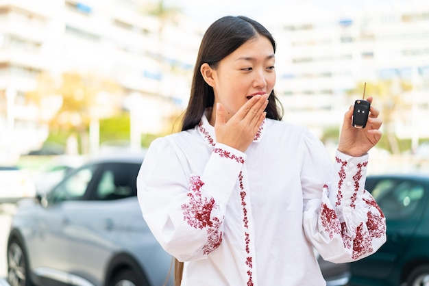 Joven china sosteniendo las llaves del auto al aire libre con sorpresa y expresión facial conmocionada