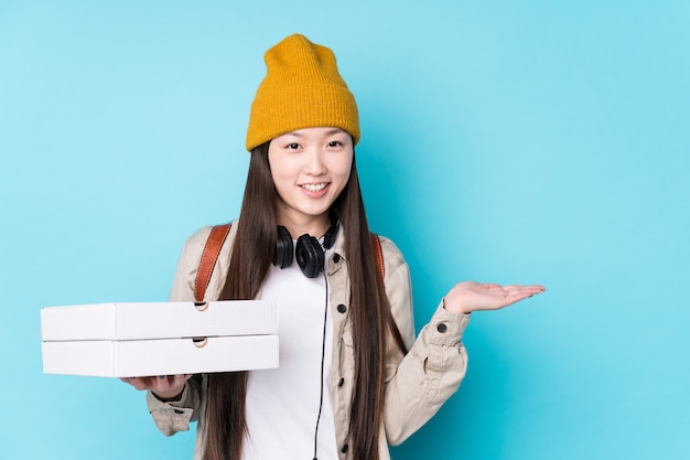 Joven China sosteniendo pizzas aisladas mostrando un espacio de copia en una palma y sosteniendo otra mano en la cintura.