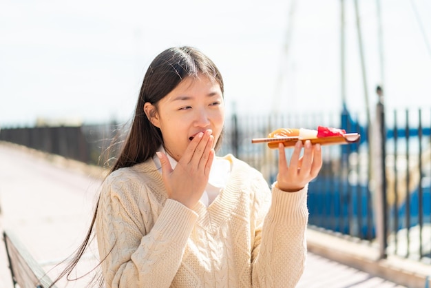 Joven china sosteniendo sashimi al aire libre con sorpresa y expresión facial conmocionada