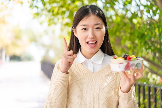 Joven china sosteniendo un tazón de fruta al aire libre señalando una gran idea
