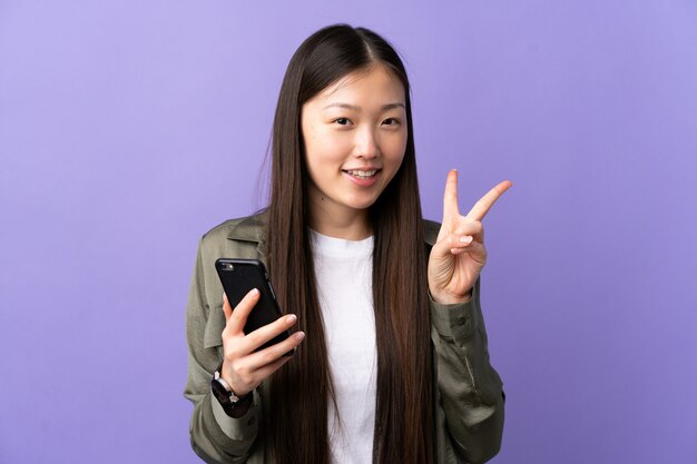 Joven china con teléfono móvil sobre pared púrpura aislado sonriendo y mostrando el signo de la victoria