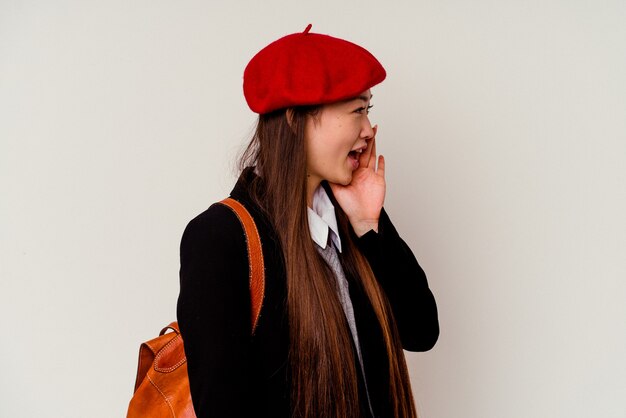 Joven China vistiendo un uniforme escolar aislado en la pared blanca gritando y sosteniendo la palma cerca de la boca abierta.