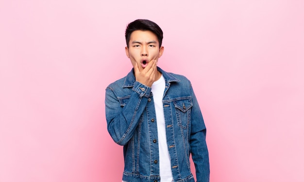 Joven chino con la boca y los ojos bien abiertos y la mano en la barbilla, sintiéndose desagradablemente sorprendido, diciendo qué o wow contra la pared de color plano