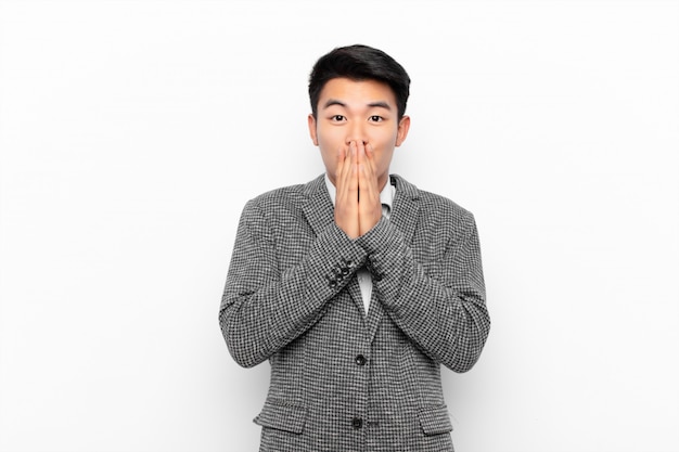 Joven chino feliz y emocionado, sorprendido y asombrado con la boca con las manos, riendo con una expresión linda en la pared de color plano