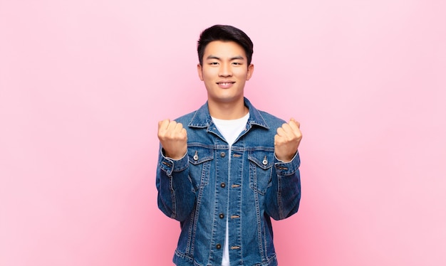 Joven chino gritando triunfante, riendo y sintiéndose feliz y emocionado mientras celebra el éxito contra la pared de color plano