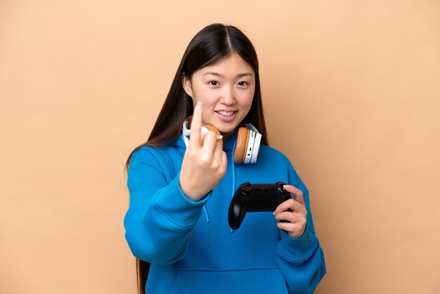 Joven chino jugando con un controlador de videojuegos aislado en un fondo beige haciendo el gesto de venir